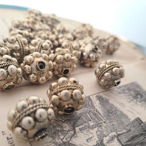 May include: A collection of silver-coloured spherical beads with a textured, dotted surface. Each bead features a small hole and a twisted rope-like band. The beads are arranged on a vintage paper background, creating a tactile contrast.