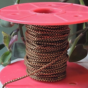 May include: A spool of bronze-colored chain sits atop a red plastic reel. The chain is tightly wound, showcasing a square-link design. The background includes green foliage, providing a natural contrast to the metallic chain.