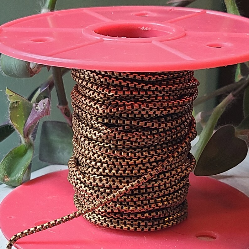 May include: A spool of bronze-colored chain sits atop a red plastic reel. The chain is tightly wound, showcasing a square-link design. The background includes green foliage, providing a natural contrast to the metallic chain.