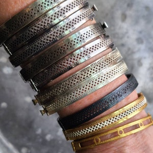 May include: A stack of silver bracelets with a delicate, patterned design. The bracelets are layered on a person's wrist, with a black leather bracelet and a gold bracelet with a white enamel design on top.