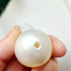 May include: A close-up of a large, round, off-white pearl bead with a hole drilled through it. The bead has a smooth, lustrous surface and is held by a person's fingers. The bead is likely for jewelry making.