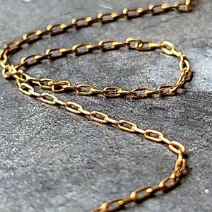May include: A close-up of a gold chain necklace with a delicate, oval-shaped link design. The chain is lying on a gray textured surface.