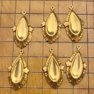 May include: Six gold-colored teardrop-shaped pendants. Each pendant has a central teardrop shape with a decorative border and a small loop at the top for hanging. The pendants are arranged on a wooden surface.