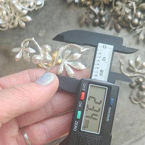 May include: A silver metal vine with leaves and berries, measured with a digital caliper showing a measurement of 23.4 mm.