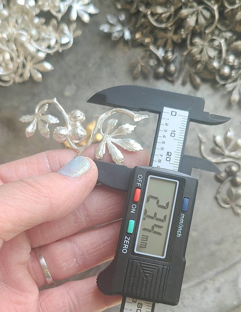 May include: A silver metal vine with leaves and berries, measured with a digital caliper showing a measurement of 23.4 mm.