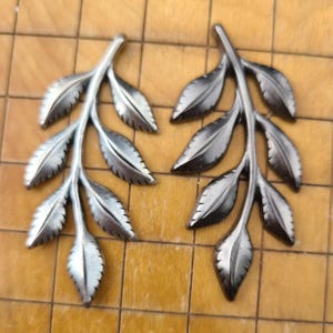 May include: Two silver-colored leaf branch ornaments. Each branch has multiple detailed leaves. The leaves have a textured appearance, and the branches are slightly curved. The ornaments are displayed on a light brown, tiled surface.