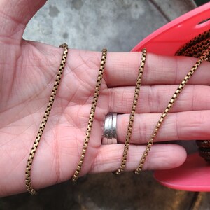 May include: A gold-colored box chain necklace. The chain is made of small, rectangular links. The necklace is displayed on a pink spool. The chain is a warm, antique gold color.