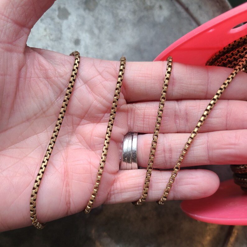 May include: A gold-colored box chain necklace. The chain is made of small, rectangular links. The necklace is displayed on a pink spool. The chain is a warm, antique gold color.