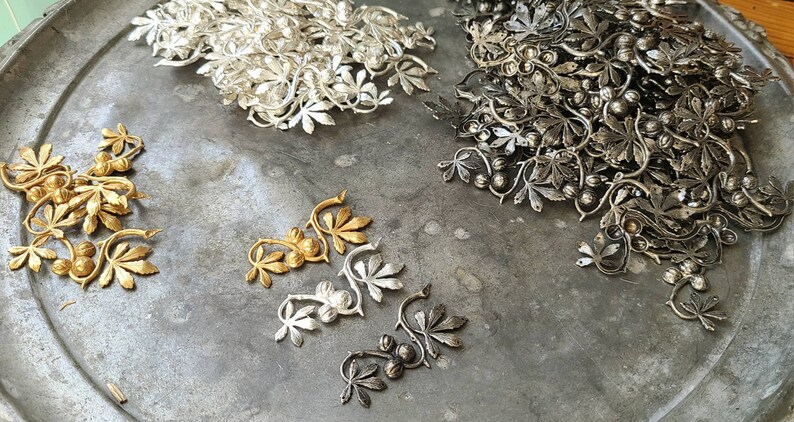 May include: A tray of metal charms in the shape of leaves and berries. The charms are in three different colours: gold, silver, and black.
