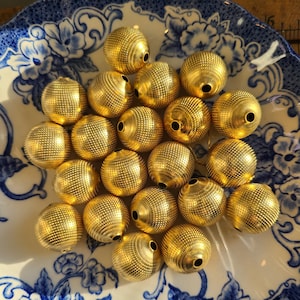 May include: A selection of gold-coloured, textured beads with small holes. The spherical beads feature a detailed, dotted pattern across their surface. They are arranged on a white and blue floral patterned plate.