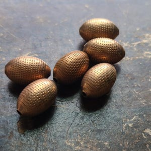 May include: Six gold oval-shaped beads with a textured surface. The beads are arranged in a semi-circle on a gray surface.