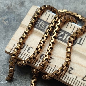 May include: Close-up of a gold-colored box chain necklace. The chain is draped over a wooden ruler, showing the intricate square links. The ruler's markings are visible, adding a sense of scale to the jewelry.
