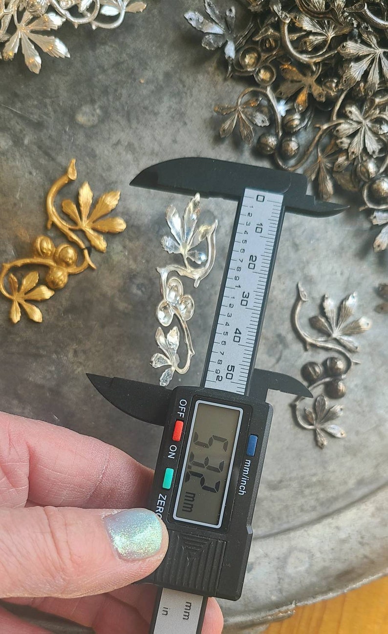 May include: A silver metal leaf design with a digital caliper measuring 6.73 cm. The caliper is black with a digital display showing the measurement in millimetres and inches.