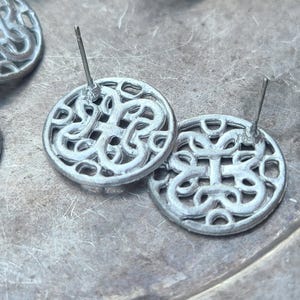 May include: A pair of silver earrings with a circular design featuring a Celtic knot pattern. The earrings have a post back closure.