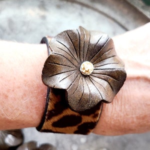 May include: A brown leather cuff bracelet with a large flower design and a gold-toned rhinestone in the center. The bracelet is made of leopard print fabric.