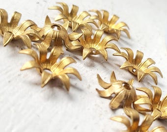 May include: Close-up of several gold-colored metal flower-shaped components. Each component has multiple petals and a central hole. The components are scattered on a white surface, suggesting a craft or jewelry-making theme.
