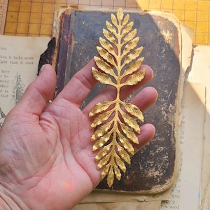 May include: A gold-colored metal fern leaf with a detailed design. The leaf is held in a hand against a brown book.