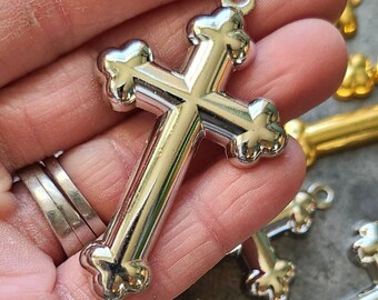 May include: Silver-colored metal cross with decorative ends and a loop at the top for hanging. The cross is held in a hand, with other crosses in the background. The crosses have a shiny, reflective surface.