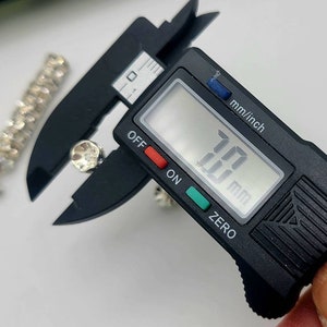 May include: A black digital caliper measuring a small, clear rhinestone. The caliper reads 10.0 mm.