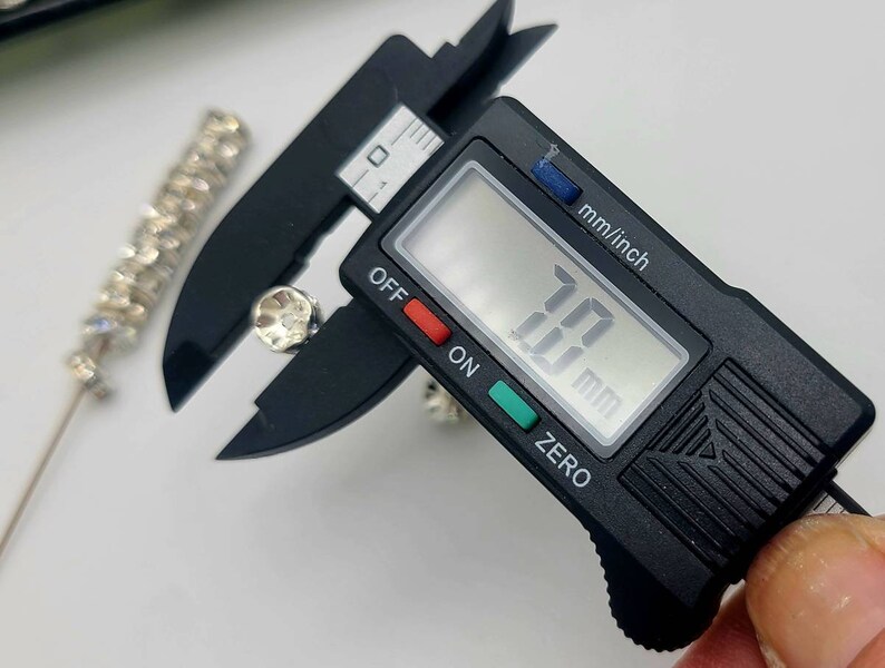 May include: A black digital caliper measuring a small, clear rhinestone. The caliper reads 10.0 mm.