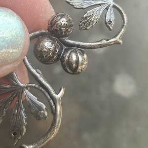 May include: A silver-toned metal charm featuring a stylised leaf with three round berries. The charm is designed to be attached to a necklace or bracelet.
