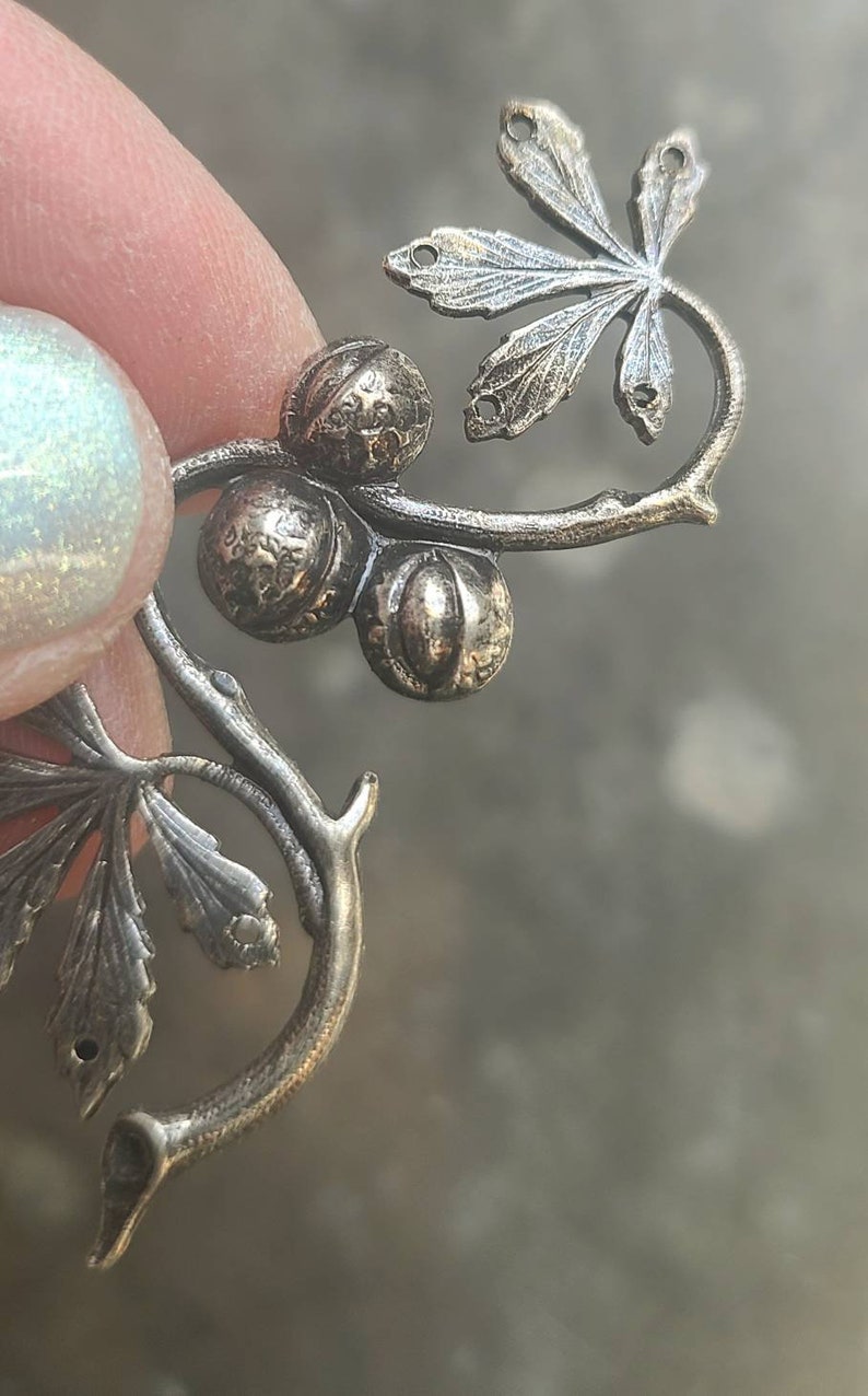 May include: A silver-toned metal charm featuring a stylised leaf with three round berries. The charm is designed to be attached to a necklace or bracelet.