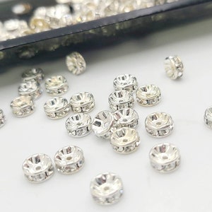 May include: A close-up of a group of silver-toned metal beads with clear rhinestones. The beads are scattered on a white surface.
