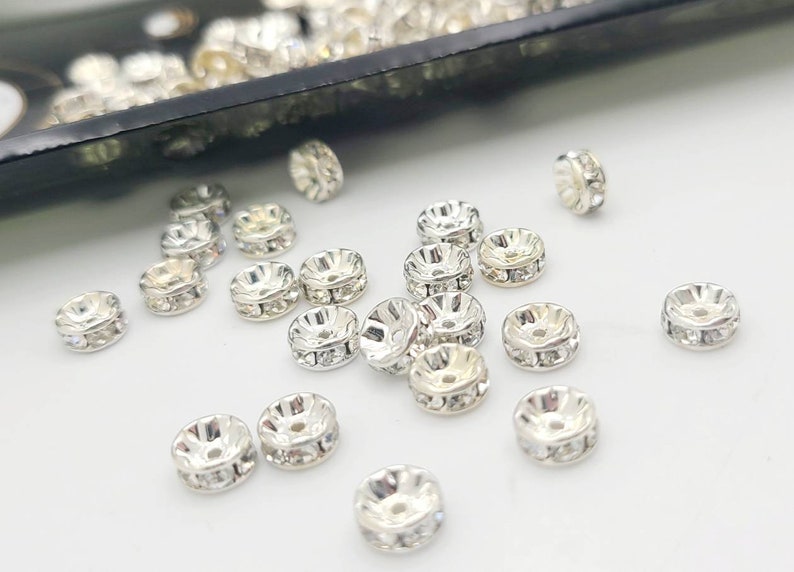 May include: A close-up of a group of silver-toned metal beads with clear rhinestones. The beads are scattered on a white surface.