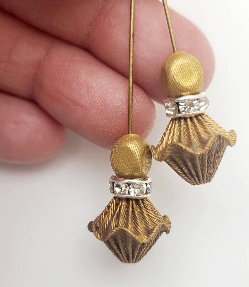 May include: A pair of gold-toned earrings with a dangling, textured, flower-shaped design. Each earring features a small, round bead and a row of clear rhinestones.