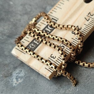 May include: A gold-colored box chain necklace is draped over a wooden ruler. The chain is made of small, square links. The ruler shows measurements in inches. The necklace is approximately 3 inches long.