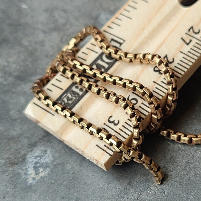 May include: A gold-colored box chain necklace is draped over a wooden ruler. The chain is made of small, square links. The ruler shows measurements in inches. The necklace is approximately 3 inches long.