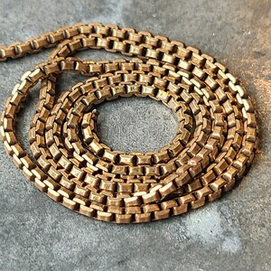 May include: A gold-colored box chain necklace, coiled on a gray surface. The chain is made of small, rectangular links, creating a textured appearance. The necklace is a classic jewelry piece.