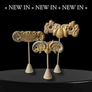 May include: Three antique brass jewellery display stands with ornate designs. Each stand features a decorative top, a slender metal rod, and a burlap-covered cone base. The text "+ NEW IN + NEW IN + NEW IN +" is at the top.