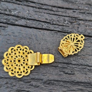 May include: Two ornate, gold-colored clasps. One is a circular shape with a detailed, openwork design. The other is an oval shape with a similar openwork pattern. Both clasps have a rectangular clasp mechanism.