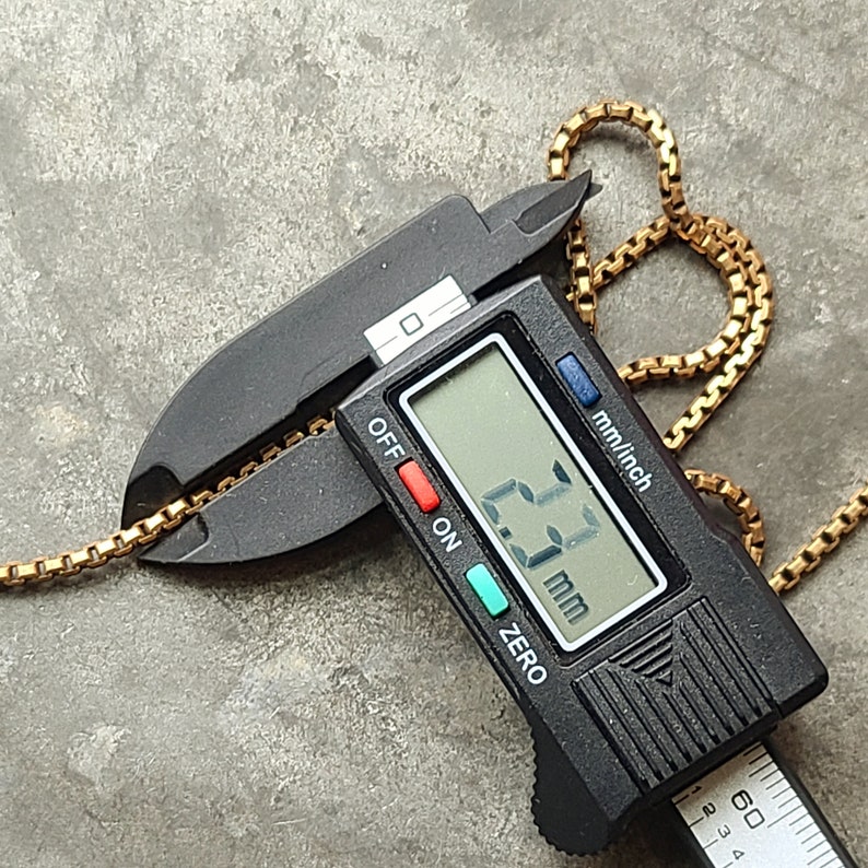 May include: A gold-colored chain is measured with a black digital caliper. The caliper displays a measurement of 2.3 mm. The chain is made of small, square links.