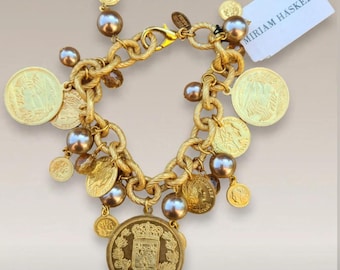 Peut inclure: Un bracelet à breloques doré composé d'une chaîne torsadée épaisse, de multiples médailles métalliques rondes avec des motifs en relief et de perles sphériques marron lustrées. Une étiquette rectangulaire blanche porte le texte MIRIAM HASKELL.