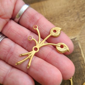 May include: Gold-toned metal charm with a stylized leaf design. The charm has three leaves and a decorative stem with a loop at the top for attaching to a necklace or bracelet.
