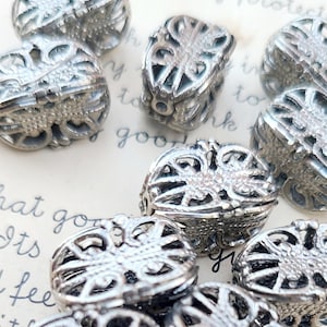May include: Close-up of several silver-colored filigree beads. The beads have an ornate, openwork design with a floral or scroll-like pattern. The beads are scattered on a light-colored surface with handwritten text.