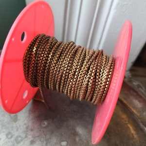 May include: A spool of gold-colored chain is wrapped around a bright pink plastic reel. The chain has a box link design. The reel has a hole on each side. The background is out of focus.