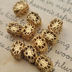 May include: A collection of gold-colored filigree beads. The beads have an openwork design with intricate patterns. The beads are oval-shaped and appear to be made of metal. They are scattered across a light-colored surface.