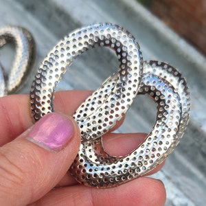 May include: Close-up of two silver-colored, oval-shaped metal links. Each link is perforated with numerous small holes, creating a textured surface. The links are intertwined, suggesting a connection or joining element. The image is well-lit, highlighting the metallic sheen.