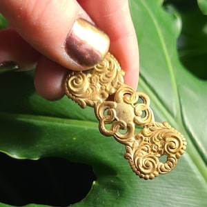 May include: A gold-colored metal decorative piece with intricate floral designs. The piece has a smooth, polished finish and is shaped like a long, narrow rectangle.