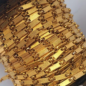 Peut inclure: Gros plan d'une bobine de chaîne dorée. La chaîne est composée de maillons rectangulaires à surface texturée, reliés par de petits maillons ronds. La chaîne est enroulée serrée sur une bobine noire, créant un motif dense et texturé.