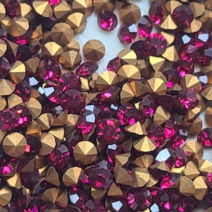 May include: A close-up image of a large quantity of small, faceted, dark red rhinestones and gold-toned metal settings. The rhinestones and settings are scattered randomly.