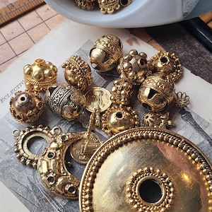 May include: A collection of gold-toned metal beads and charms, including a large round pendant with a central hole. The beads have intricate designs and some are round, while others are more irregular in shape. The charms include a small flower and a coin-like piece.