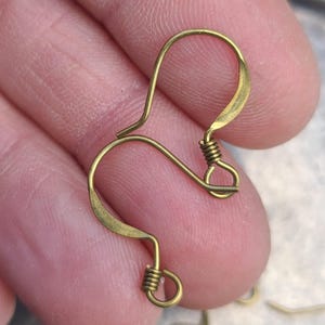 May include: Close-up of two gold-colored earring hooks. Each hook features a curved design with a looped end and a coiled wire detail. The hooks are held in a hand, showcasing their delicate craftsmanship.