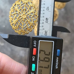 May include: A gold-colored metal decorative button with a filigree design. The button is being measured with a digital caliper, which reads 29.7 mm.