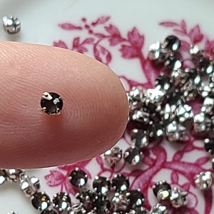 Peut inclure: Un seul strass noir sur le bout d'un doigt, entouré d'une dispersion de strass argentés sur un fond floral rose et blanc.