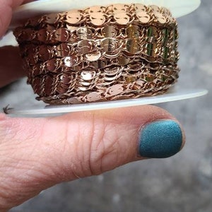 Puede incluir: Una bobina de gal&oacute;n de cadena de color oro rosa con peque&ntilde;as lentejuelas redondas. La cadena est&aacute; superpuesta en la bobina, creando un efecto decorativo.