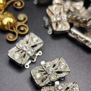 May include: Three silver-toned metal clasps with clear rhinestone embellishments. The clasps are in the shape of a bow and have a decorative design.